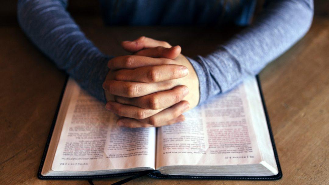 Man's folded hands with open Bible on table